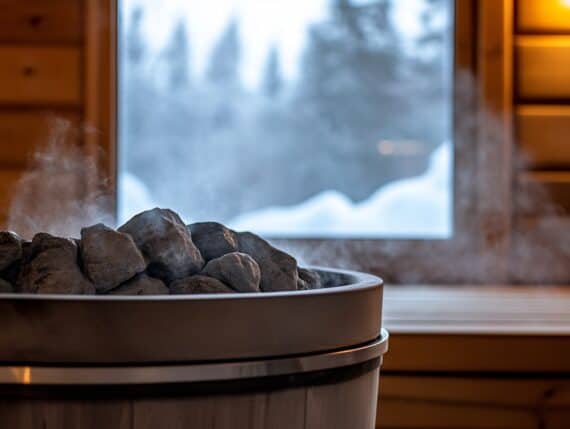 Holzsauna mit Steinofen und Blick auf verschneite Winterlandschaft durch das Fenster.